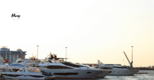 A row of luxury yachts docked at a marina during sunset, with calm water reflecting the boats and a faint Marine SEO logo in the sky above the scene.