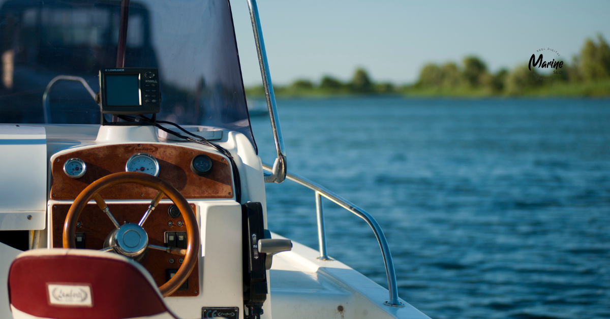 Close-up of a boat’s helm with a wooden steering wheel, gauges, and a mounted navigation device, overlooking calm blue water and a distant shoreline with trees.
