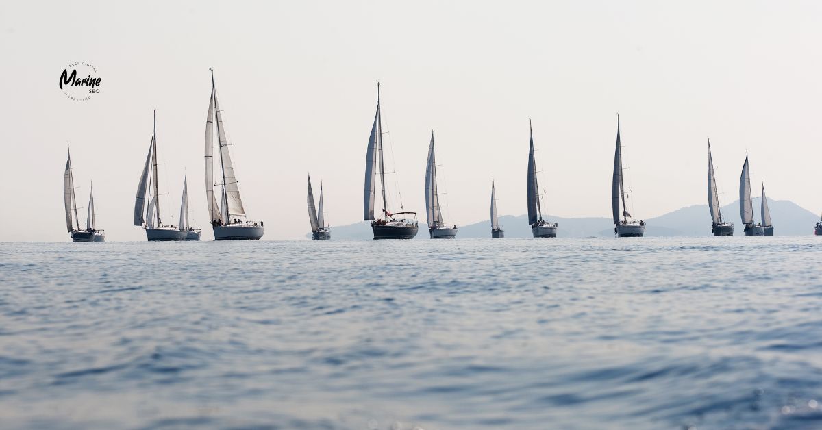 Sailboats lined up on open water during a regatta, with distant coastline in the background.