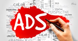 A hand holding a black pen pointing toward the word "ADS" written in large white letters inside a bright red paint splatter, surrounded by a word cloud of marketing and design terms.